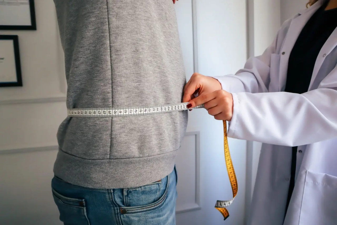 A doctor measuring the waist of a person in a grey sweater.